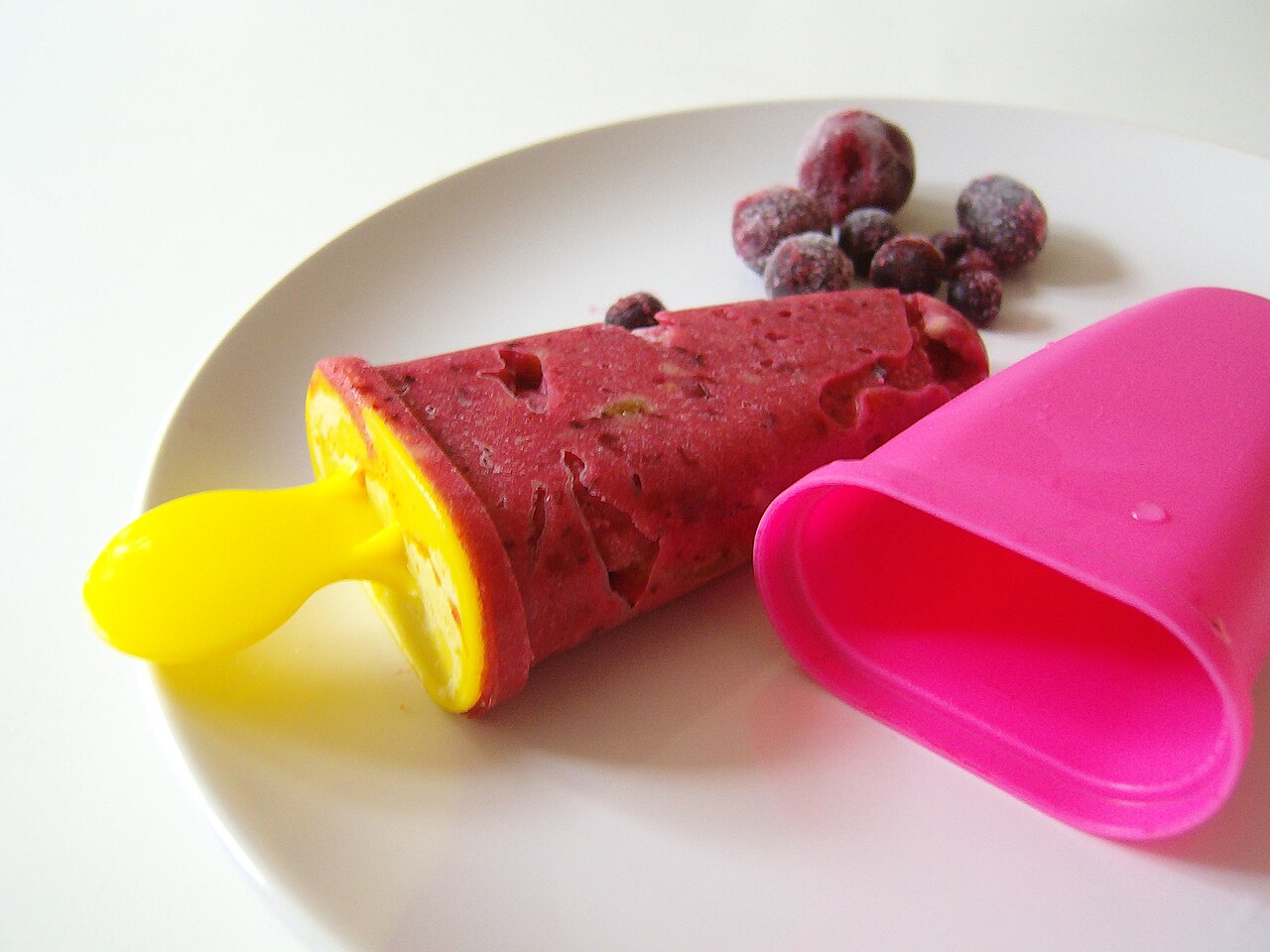 Red fruit ice pop beside frozen berries on a white plate